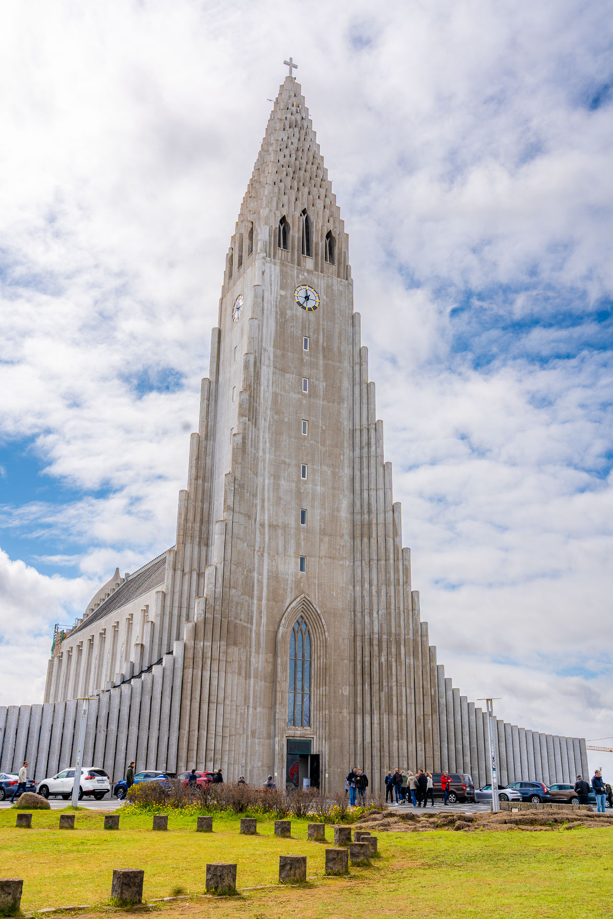 The tall concrete tower of Hallgrimskirkja, Reykjavik rises above the square with people and parked cars at its base.