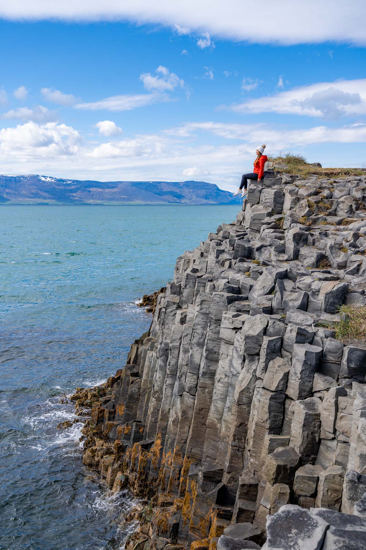 A woman in a red jacket sits on the edge of the basalt cliffs at Hofsos, Iceland above the ocean with mountains in the distance.