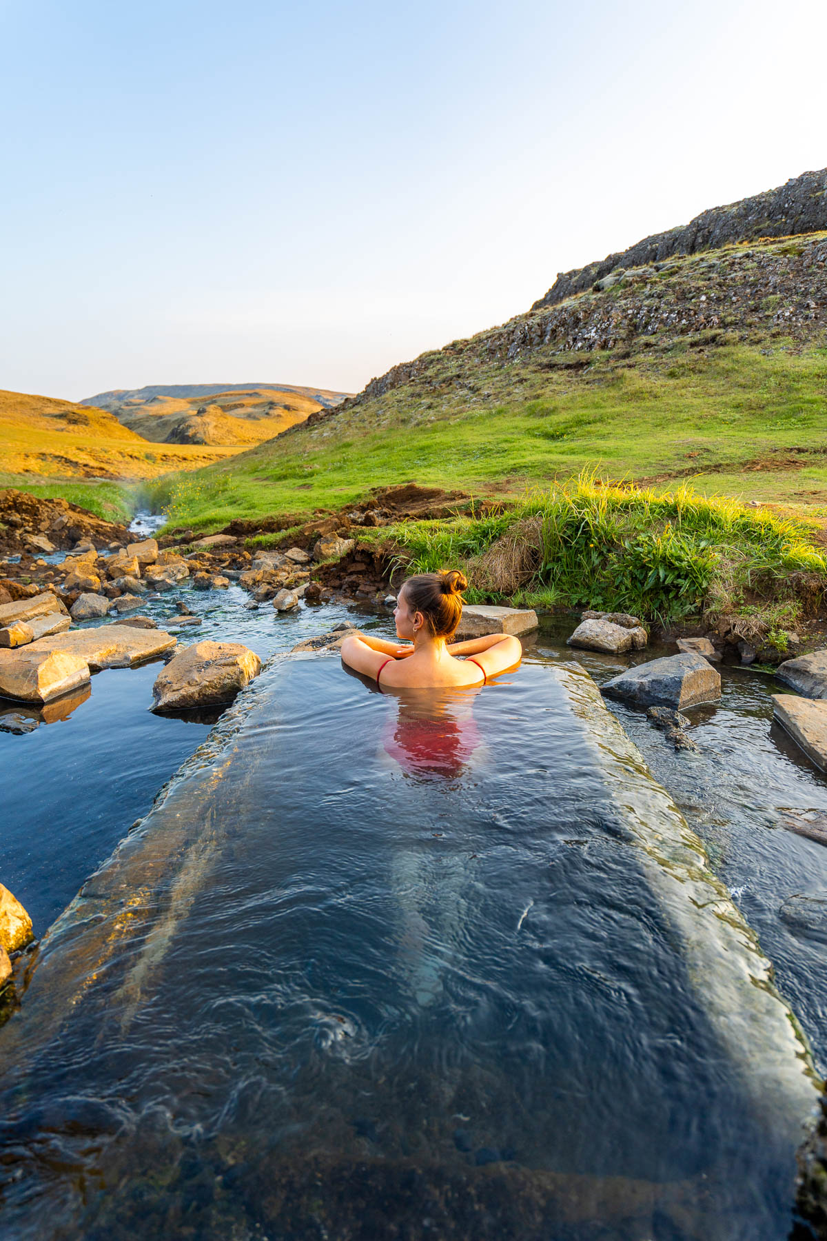 A woman relaxes in the small stone pool at Hrunalaug Hot Spring, Iceland surrounded by rocks, green hills, and a narrow stream.