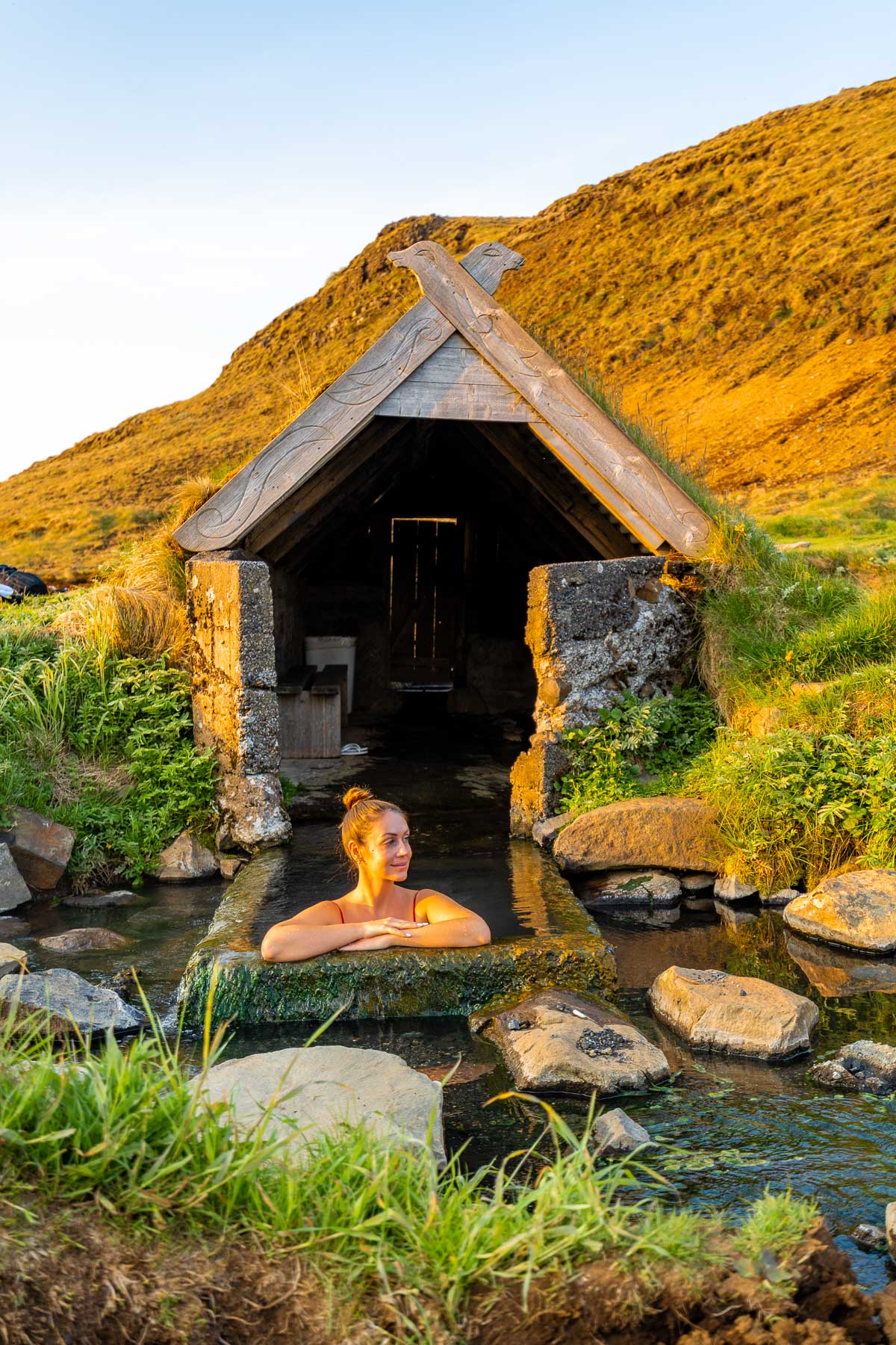 A woman leans on the edge of the small stone pool at Hrunalaug Hot Spring, Iceland with a turf-roof shelter and golden hills behind her.