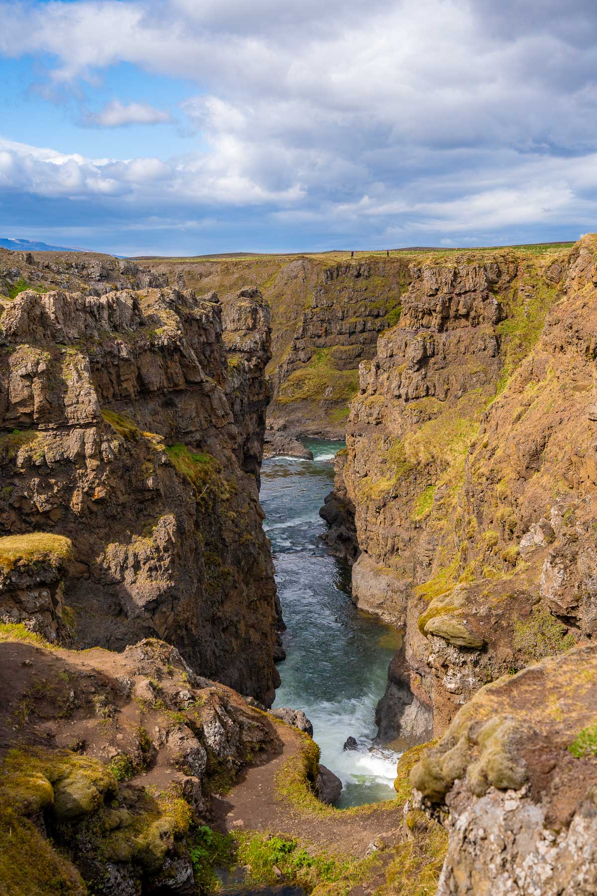 A deep crack in the landscape at Kolugljufur Canyon, Iceland reveals a fast river below, framed by rugged cliffs and uneven dirt paths along the rim.