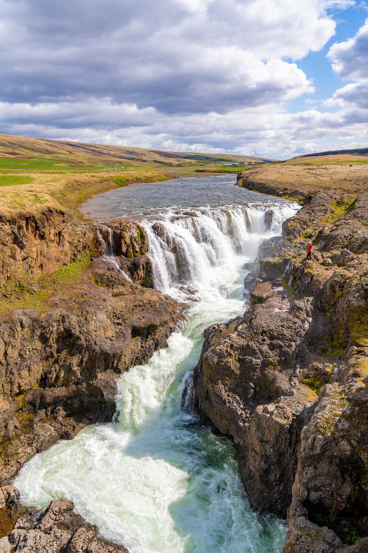 A wide view of Kolugljufur Canyon, Iceland shows the waterfall plunging into a narrow gorge with a woman in a red jacket standing on the rocky edge.