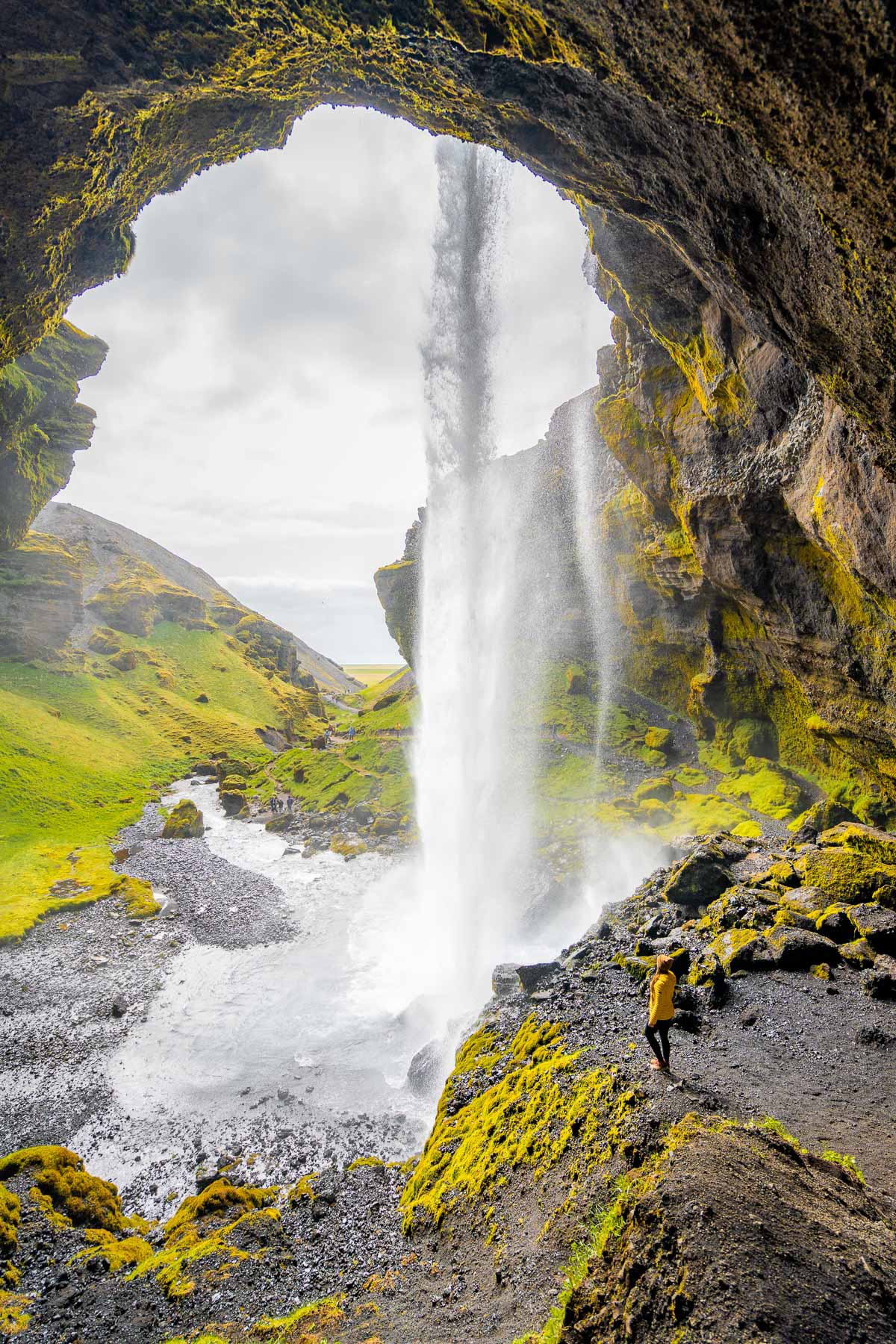 From inside a cave, Kvernufoss, Iceland drops in a single stream to the river below, framed by moss-covered rock walls and a woman in a yellow jacket on the path.