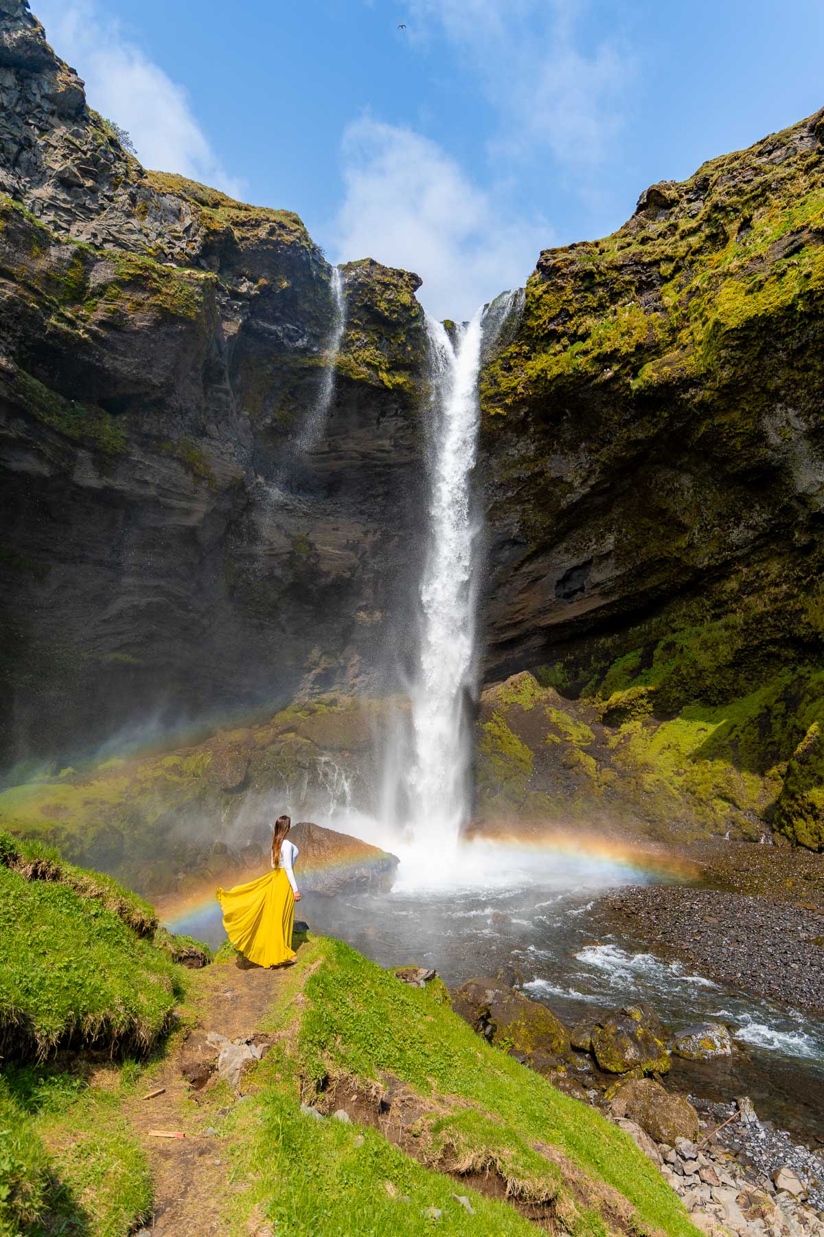 A woman in a long yellow skirt stands near the base of Kvernufoss, Iceland with mist rising and a rainbow stretching across the water.