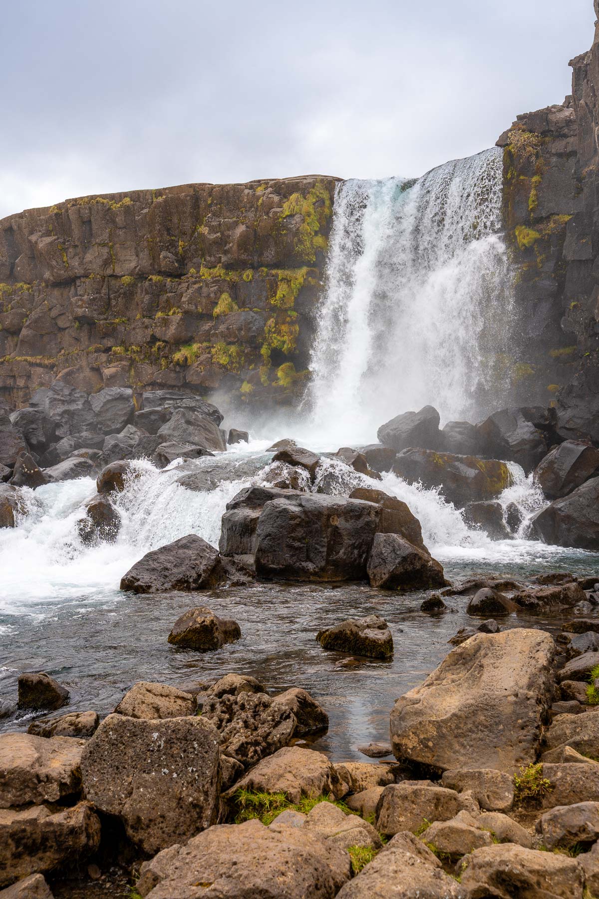 Oxararfoss in Thingvellir National Park crashes over a rocky ledge into a pool below, surrounded by large boulders and mossy cliffs.