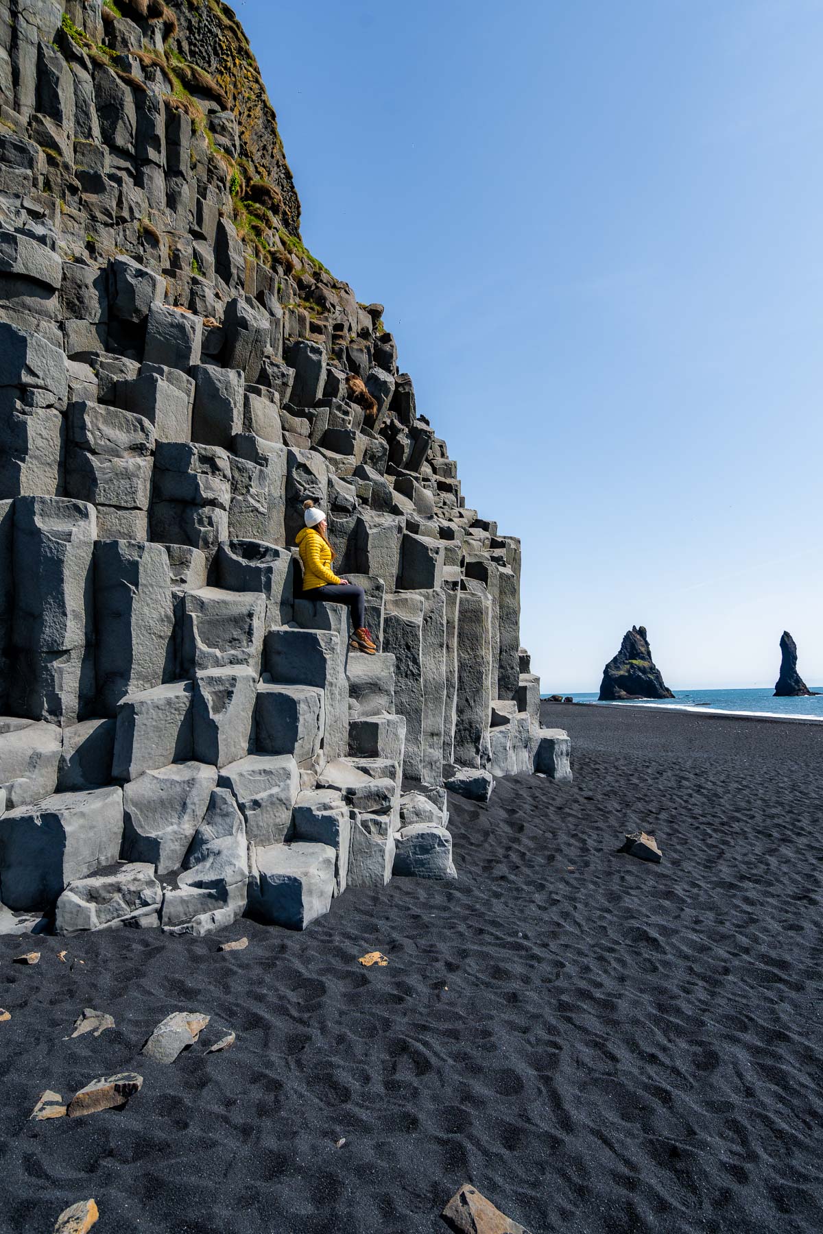 A woman in a yellow jacket sits on the basalt columns at Reynisfjara Beach, Iceland with black sand and sea stacks in the background.