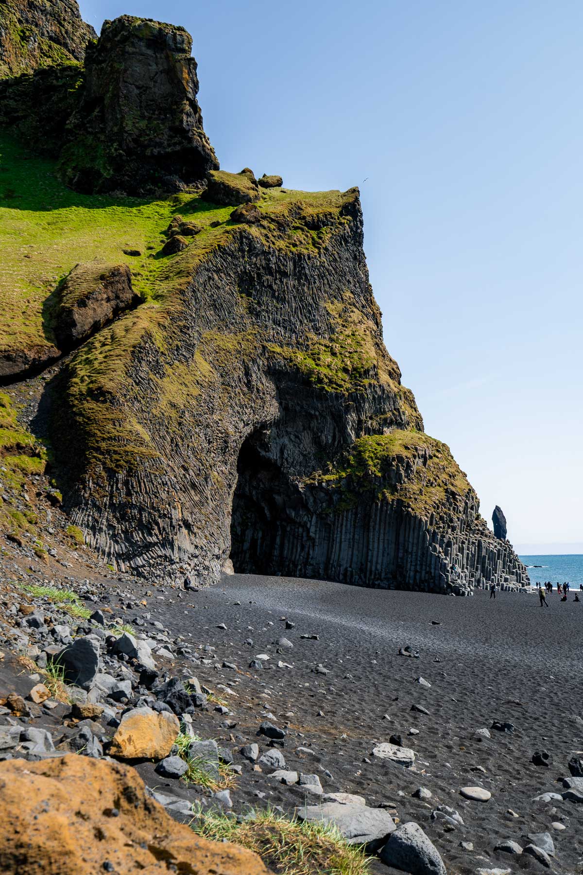 Steep cliffs and basalt columns rise above the black sand at Reynisfjara Beach, Iceland with a few people walking along the shore.