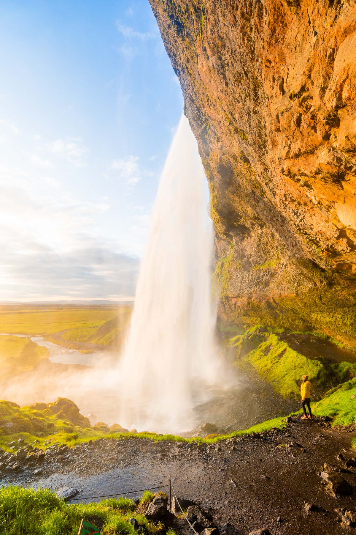 A woman in a yellow jacket stands on the path behind Seljalandsfoss, Iceland as the waterfall drops in front of her with green fields stretching into the distance.