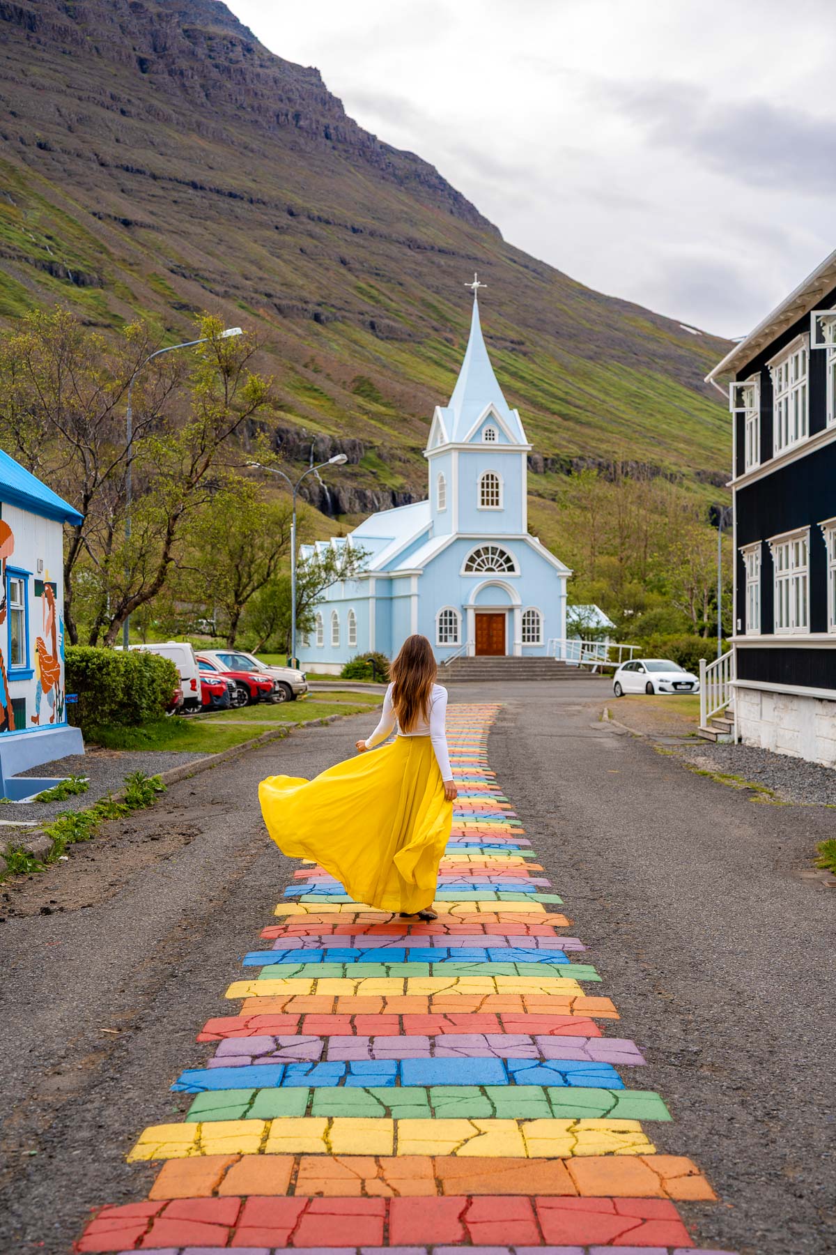 A woman in a yellow skirt walks along the rainbow street in Seydisfjordur, Iceland toward the light blue church with mountains rising behind it.