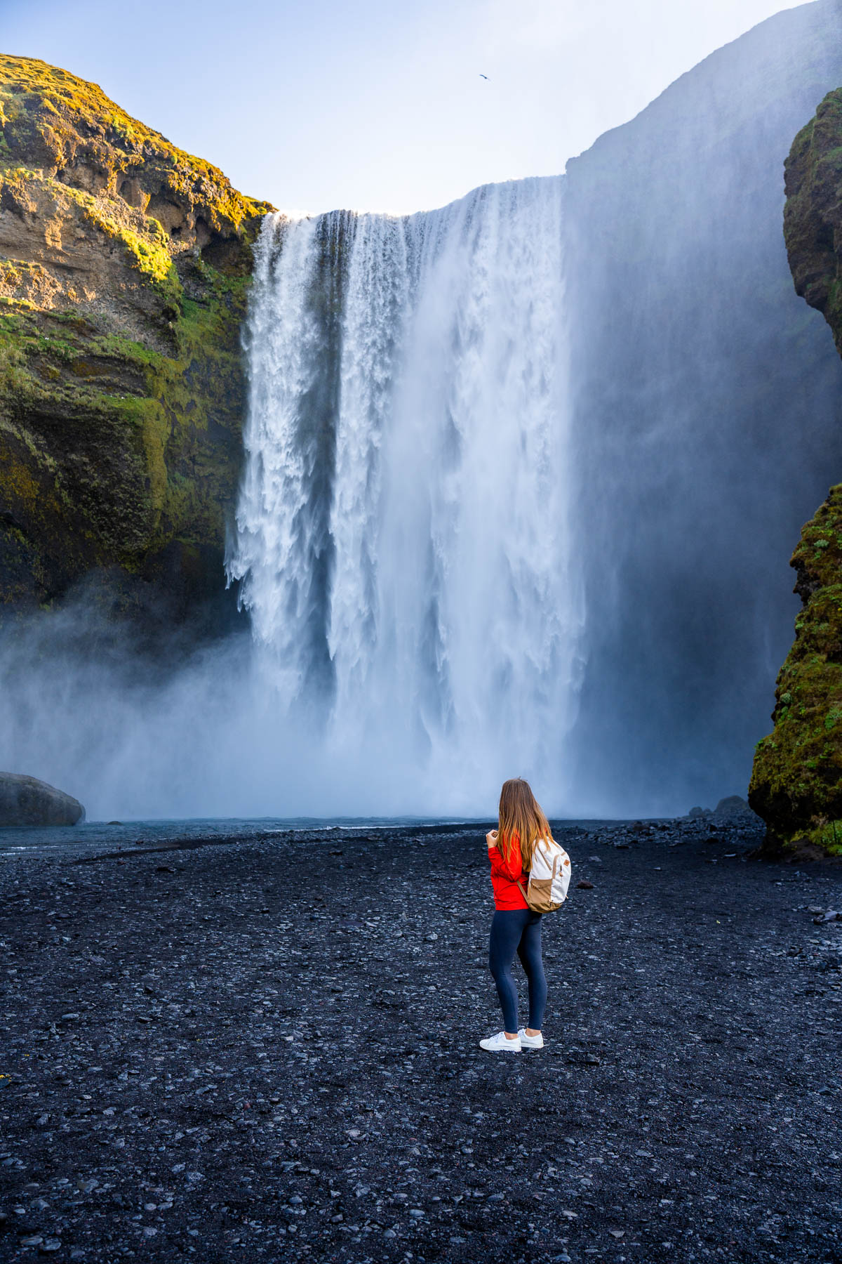A woman in a red jacket stands on the black sand in front of Skogafoss, Iceland as the wide waterfall crashes down behind her.