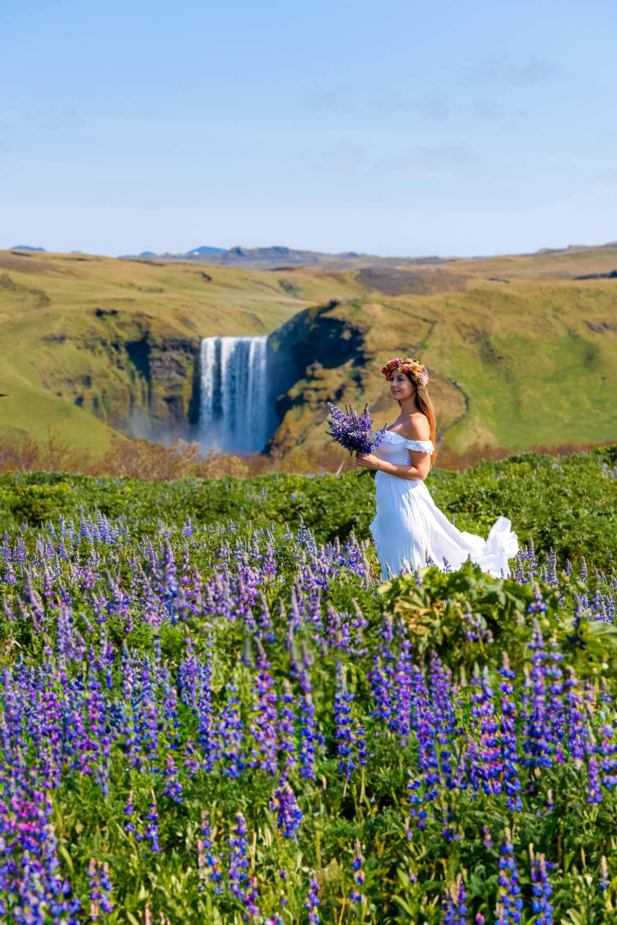 A woman in a white dress and flower crown stands in a field of purple lupins with Skogafoss, Iceland flowing in the background.