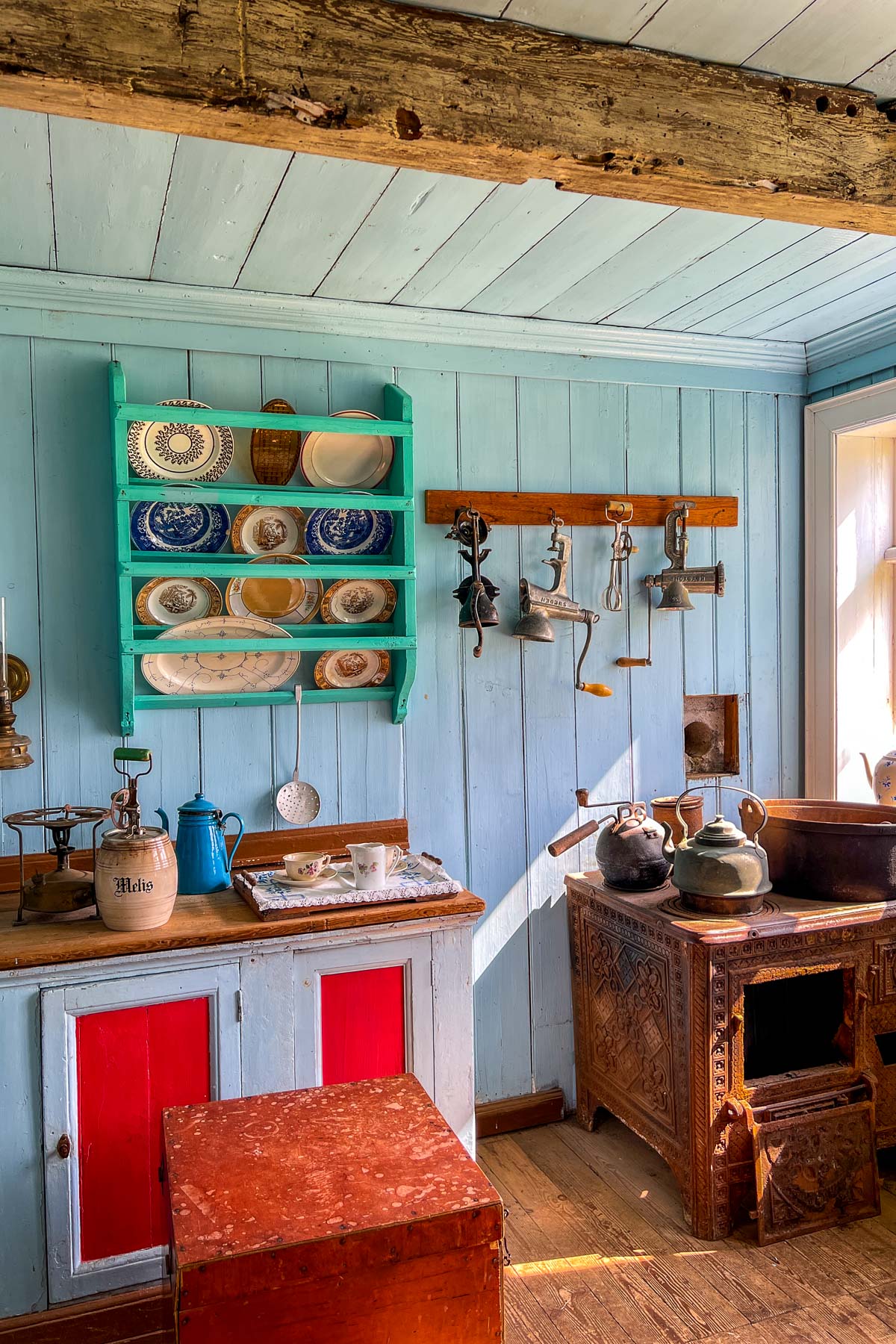 Inside Skogar Museum, Iceland a light blue wooden kitchen shows hanging metal tools, a carved stove, shelves with plates, and old kettles on display.