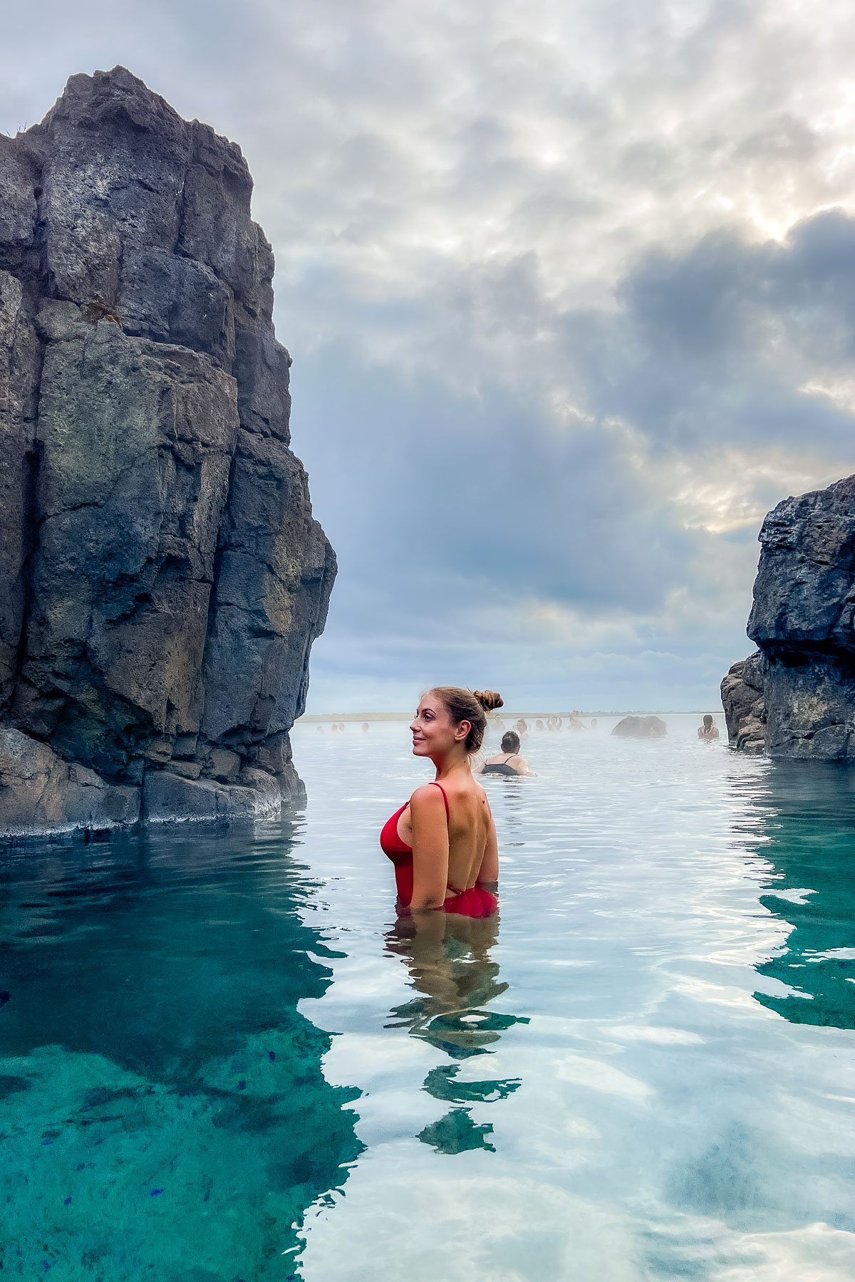 A woman in a red swimsuit stands in the blue water at Sky Lagoon, Iceland between dark rock formations with steam rising around her.
