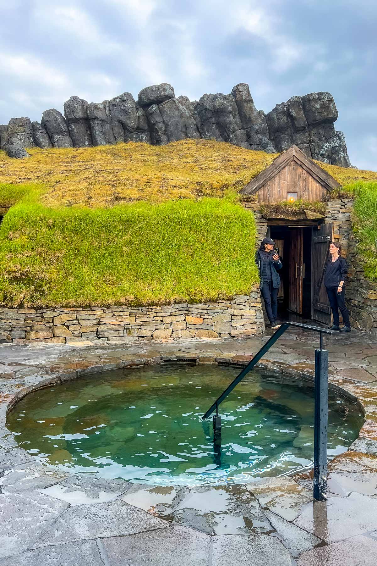 A small round hot pool sits outside a turf building at Sky Lagoon, Iceland with two staff members standing by the open wooden door and basalt cliffs behind.