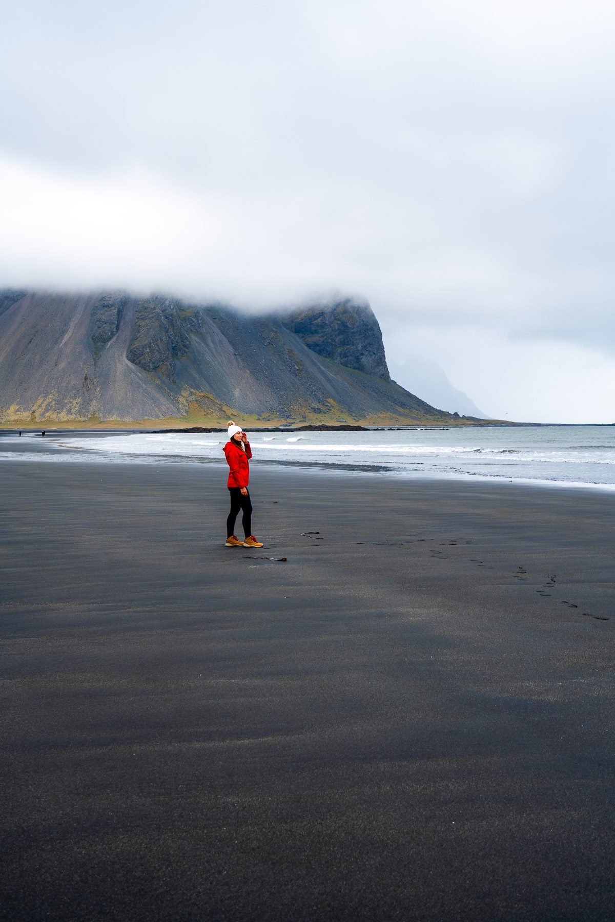 A woman in a red jacket walking along the black sand beach at Stokksnes, Iceland, with low clouds covering the peaks of Vestrahorn in the background.