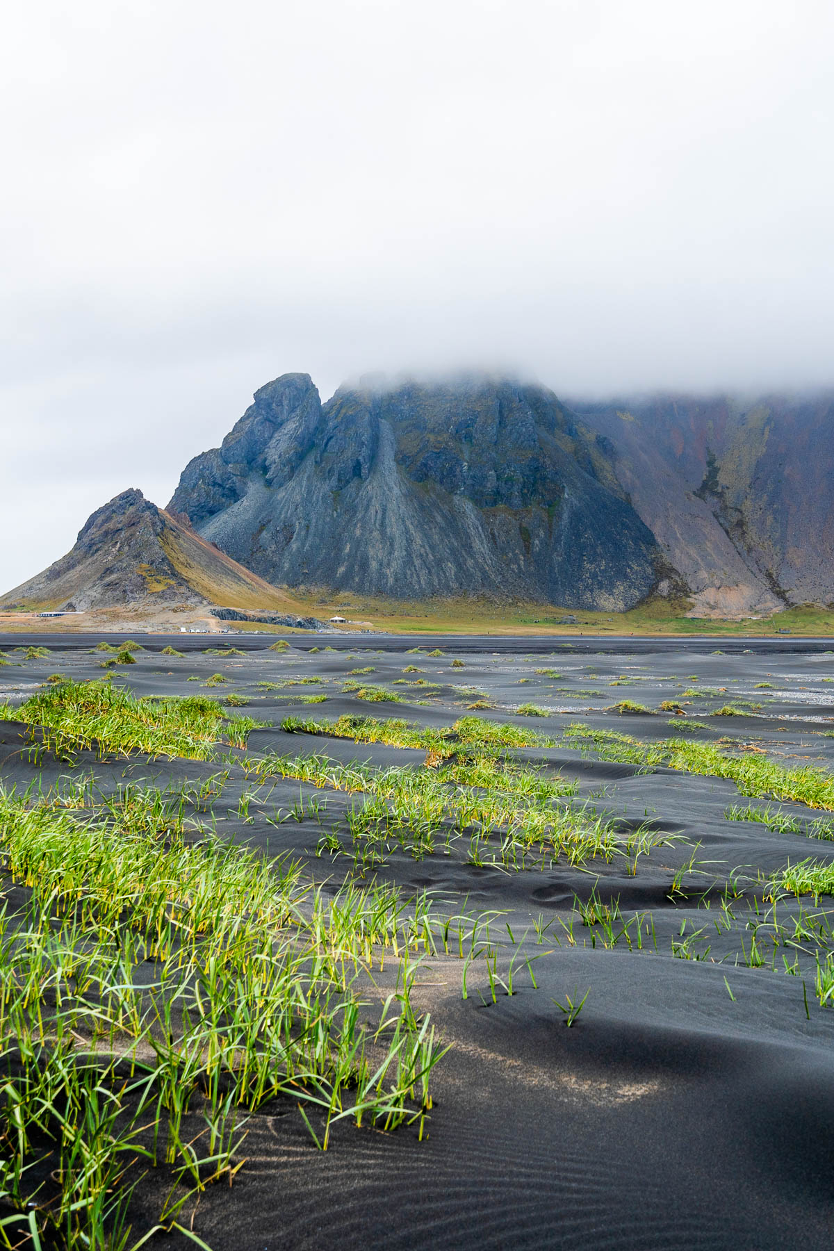 Black sand dunes with patches of green grass at Stokksnes, Iceland, leading toward the steep mountain rising under low clouds.
