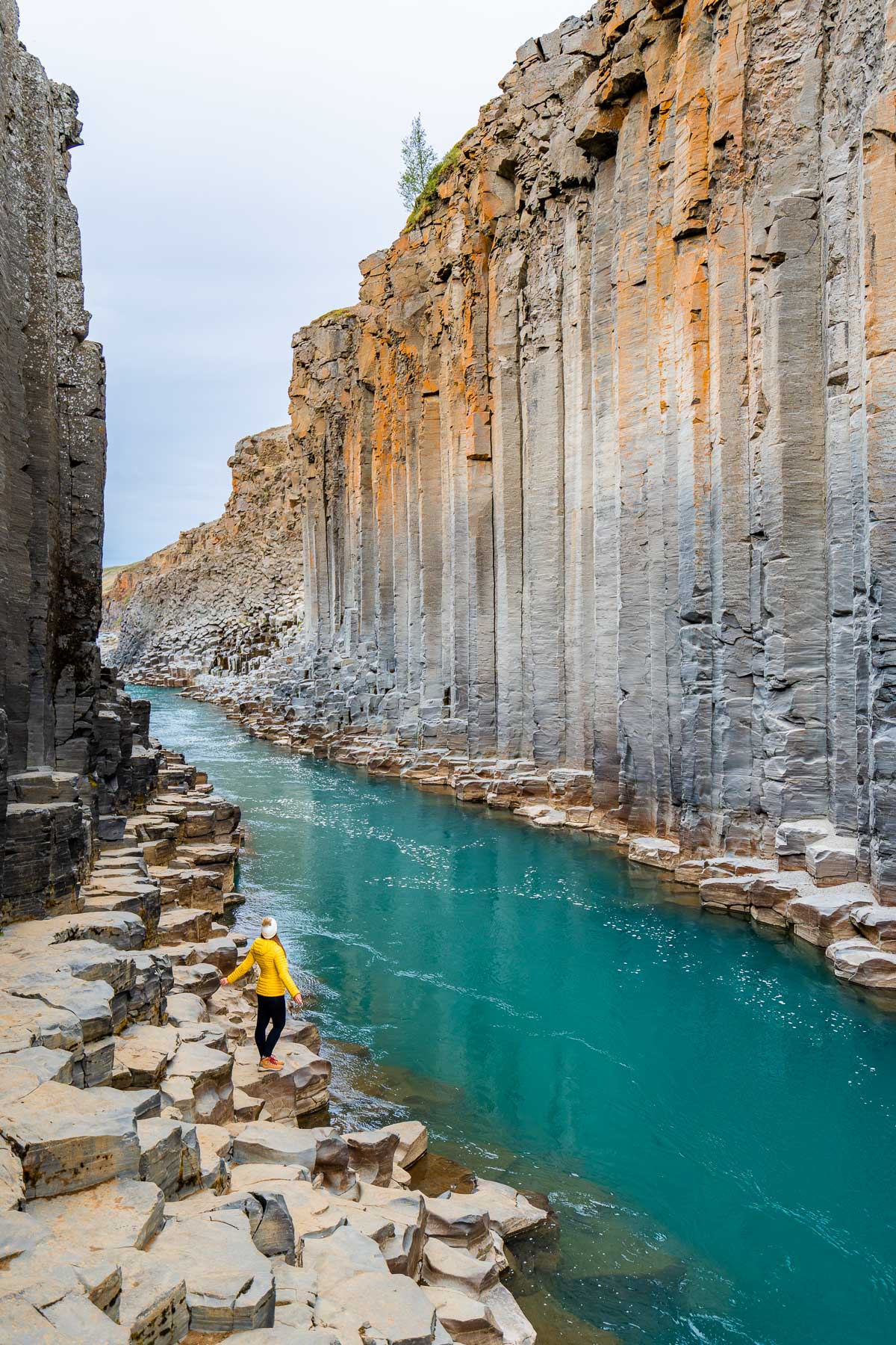 A woman in a yellow jacket standing on the rocks inside Studlagil Canyon in Iceland, surrounded by steep basalt columns and bright blue water running through the narrow gorge.