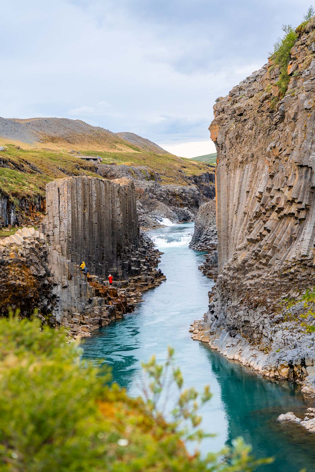 Looking down into Studlagil Canyon in Iceland where the river cuts between high basalt walls, with a few people visible on the rocky ledges near the water.