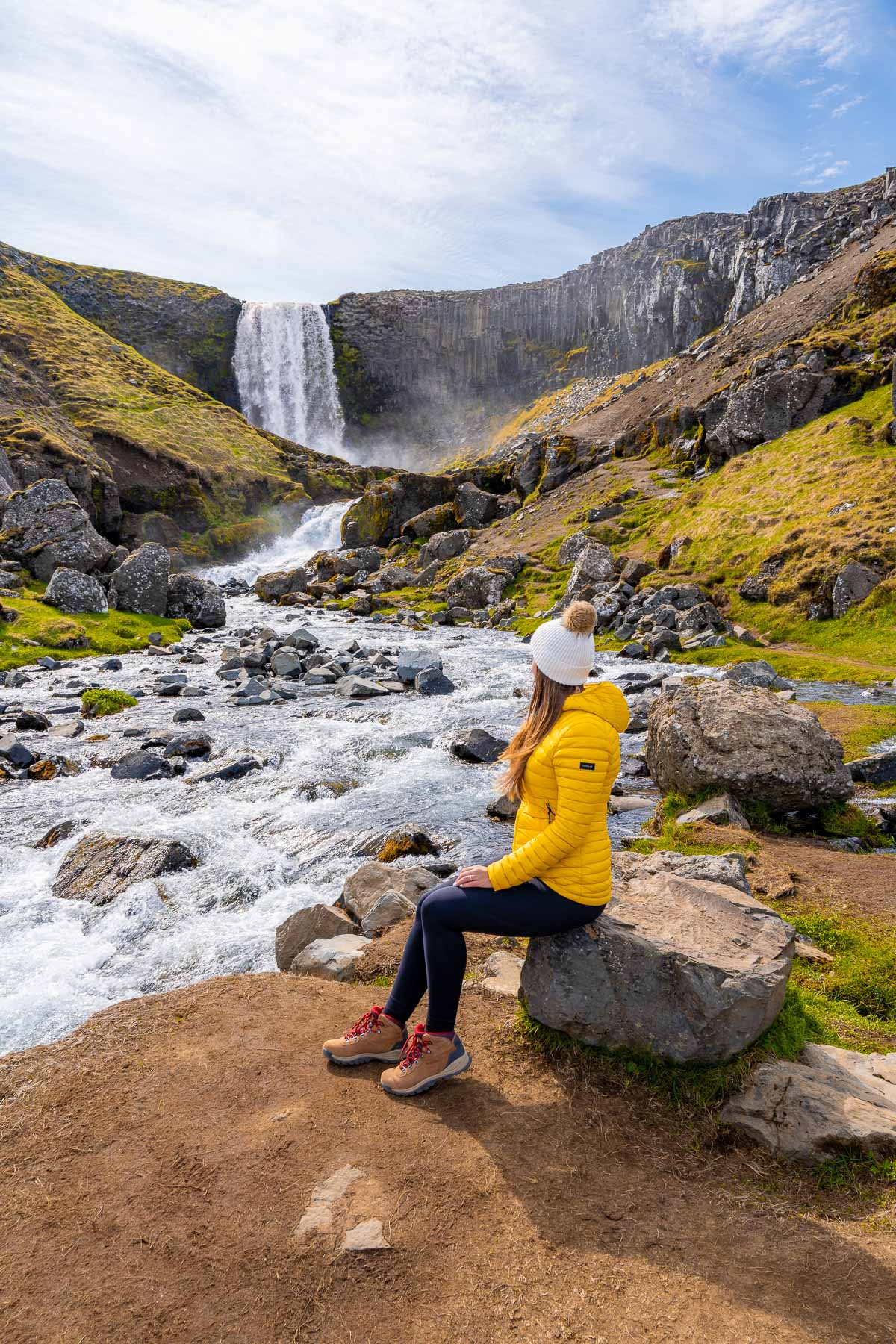 A woman in a yellow jacket and white beanie sits on a rock beside a rushing stream at Svodufoss in Iceland, looking toward the wide waterfall framed by tall basalt columns and green hills.