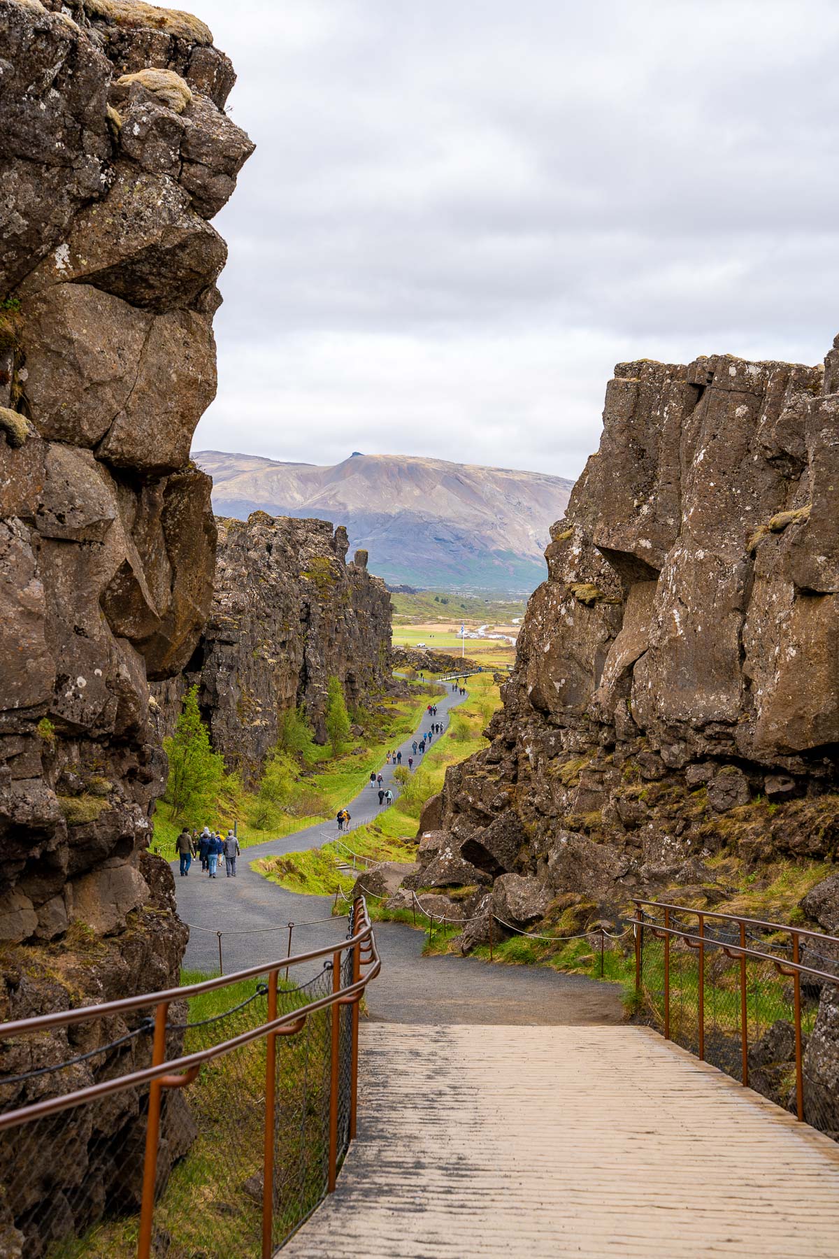 A wooden boardwalk with metal railings leads down between rocky cliffs at Thingvellir National Park in Iceland, with visitors walking along the paved path and mountains visible in the distance.