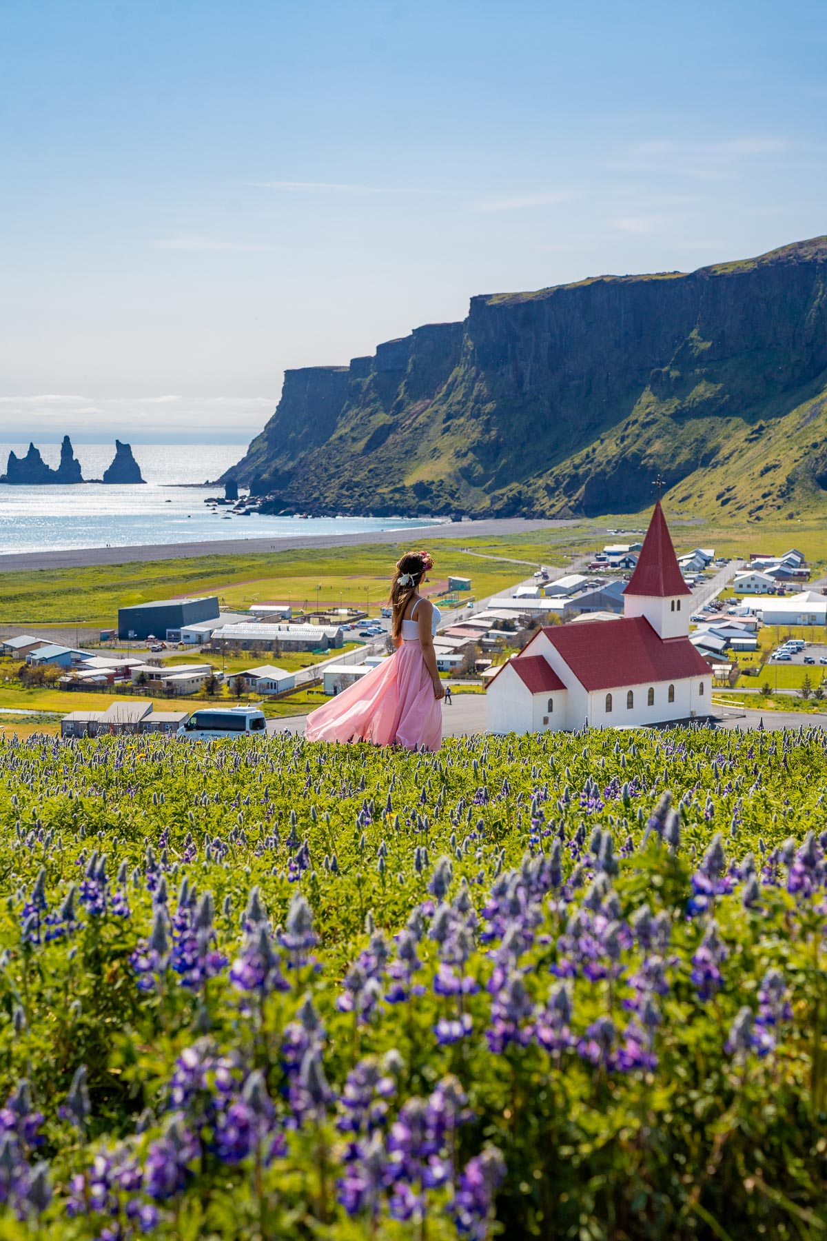 A woman in a pink dress stands in a field of purple lupines overlooking Vik i Myrdal Church, Iceland, with the red-roofed white church, the village, black sand beach, and sea stacks in the background.