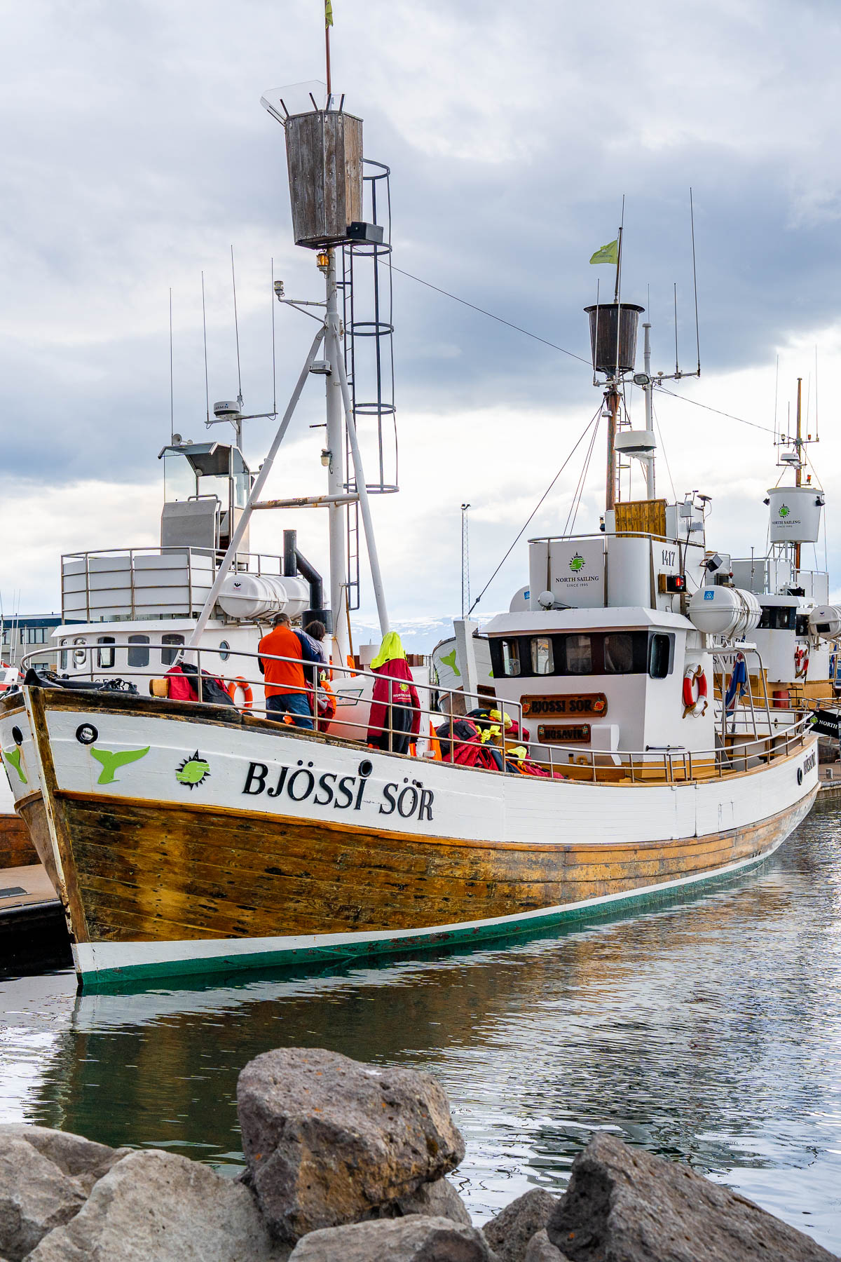 The wooden boat Bjössi Sör is docked in Husavik, Iceland, with people in colorful jackets standing on deck before a whale watching tour.